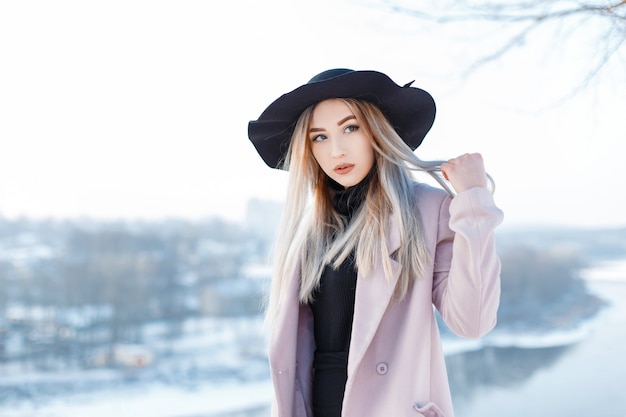 Sensual beautiful young woman with blond hair in a vintage hat in a winter pink coat in retro style is standing outside