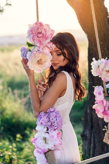 Sensual cute girl in a white dress near a wooden swing decorated with tender colored flowers in the sunset.