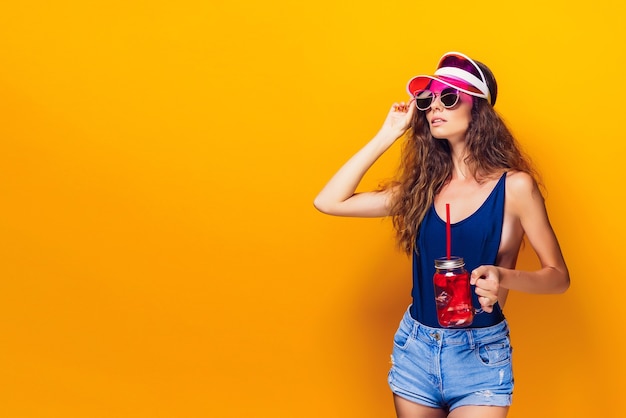 Sensual young woman in summer outfit, cap, sunglasses holding jar with fresh beverage and while standing on bright yellow