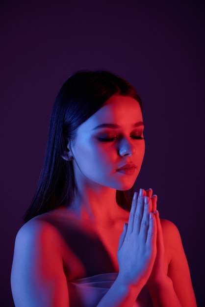 Serene calm young woman with naked shoulders meditating with closed eyes against dark blue background, neon light