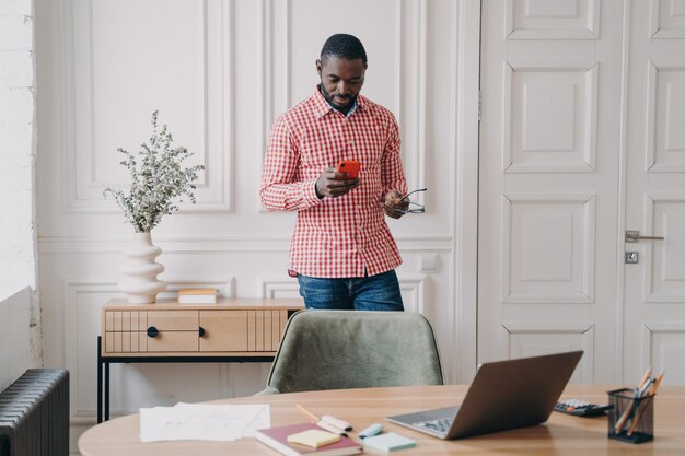 Serious African American businessman in casual attire using smartphone in office reading news chatting with clients making work calls