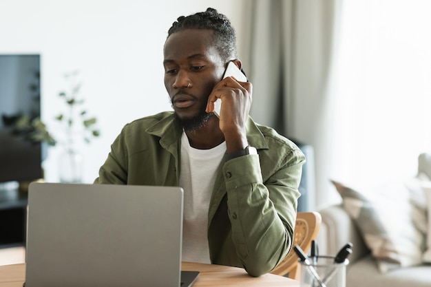 Serious african american male freelancer talking on cellphone and looking at laptop computer working online from home