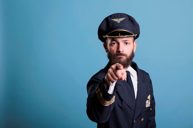 Serious airplane aviator pointing at camera with finger, aviation academy pilot training recruitment. Confident finger wearing air crew uniform and hat front view portrait