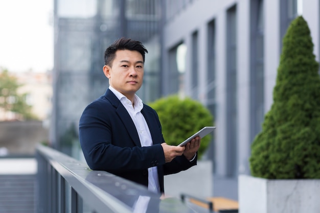 Serious asian boss reads news from tablet near modern office male businessman in business suit