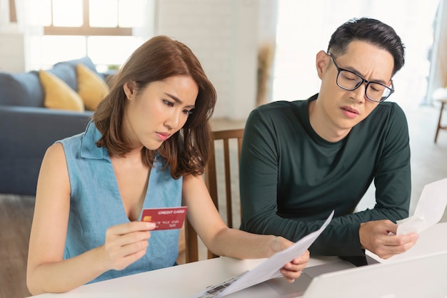 Serious Asian husband checking analyzing statement utilities bills sitting together at home.