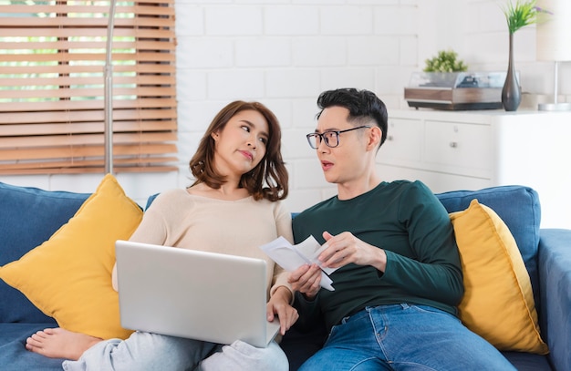 Serious Asian husband checking analyzing statement utilities bills sitting together at home.