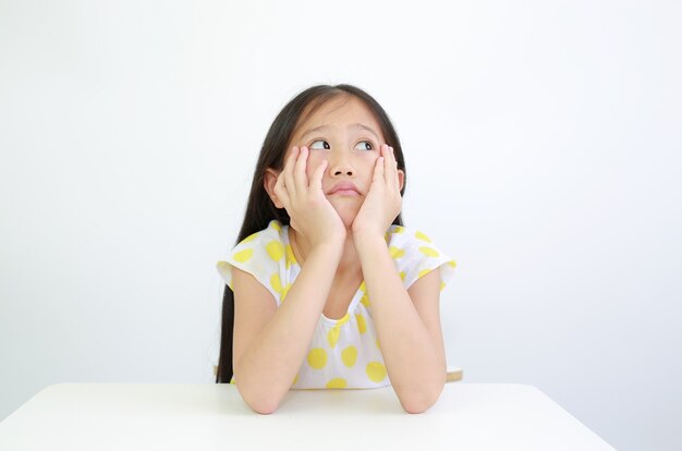 Serious Asian little child girl resting chin on hands with looking up on table over white background