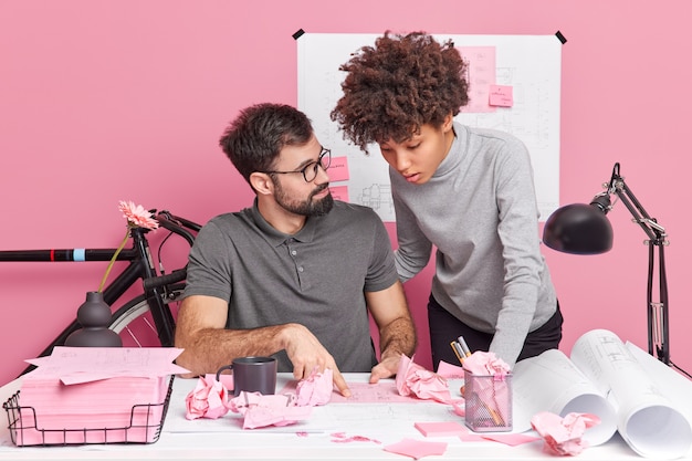 Serious attentive woman and man colleagues collaborate together pose at desktop with crumpled papers around prepare for brainstorming meeting in engineering work on project sketches of architect order