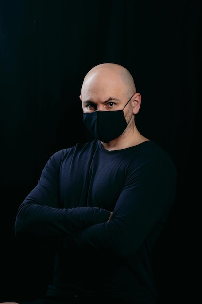 A serious bald man in a black medical mask crossed his arms over his chest Portrait of a man on a dark background