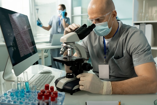 Serious bald man in protective goggles and facial mask focused on medical research sitting at desk with computer and using microscope