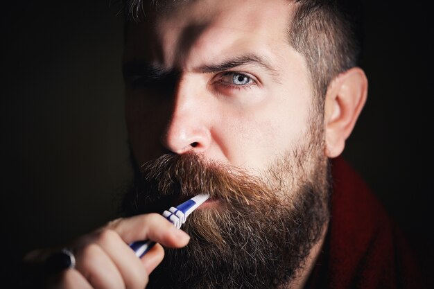 Serious bearded man in white T-shirt brushing teeth