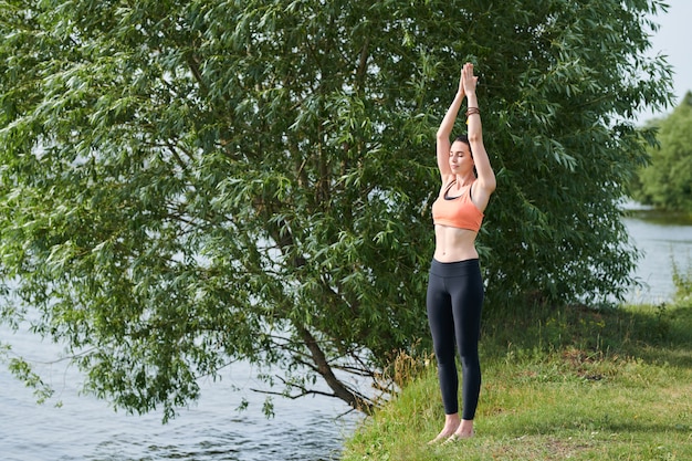 Serious beautiful young woman in sportswear standing on shore and raising hands together up while practicing yoga