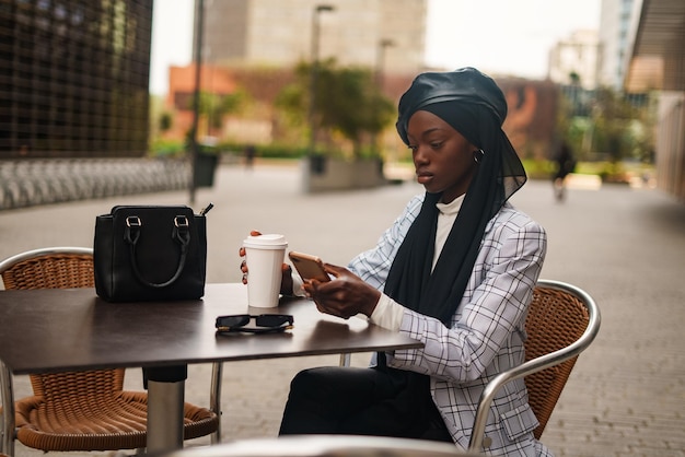 Serious black woman using smartphone at table in outdoor cafe