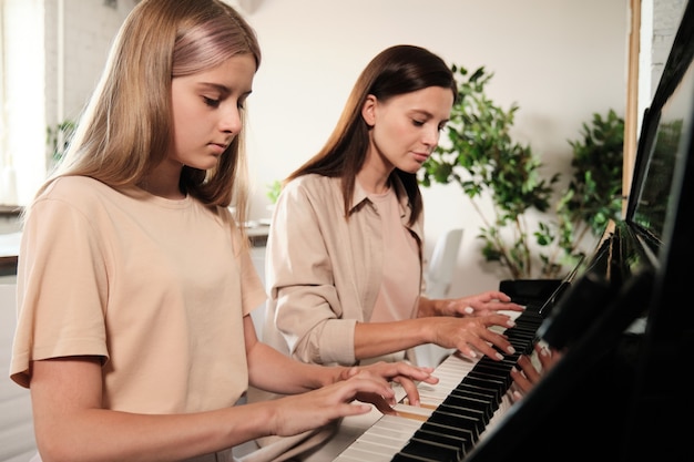 Serious blond teenage girl pushing keys of piano while training during home lesson of music with her mother sitting near by