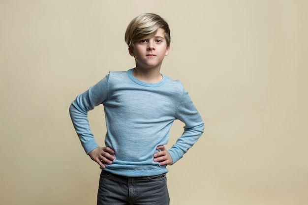 A serious boy of 910 years old stands near a yellow wall A guy in a blue sweater and gray jeans keeps his hands on his belt Confidence and positivity