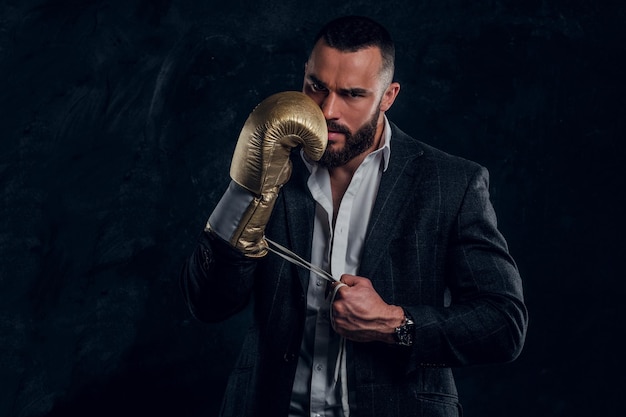 Serious brutal man in suit and golden boxing glove is posing for photographer at dark photo studio.