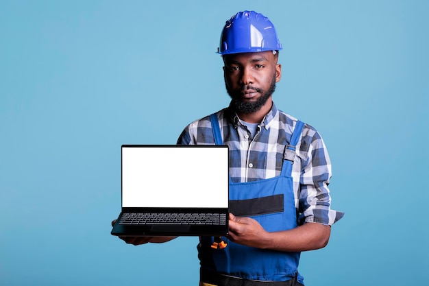 Serious builder looking at camera holding laptop with blank screen for advertisement. African american construction worker showing portable device with blank screen copy space on blue background.