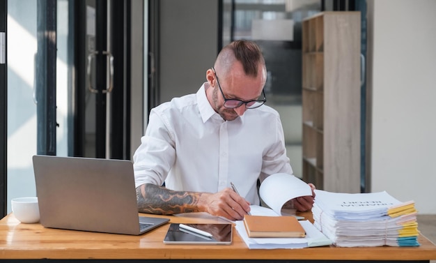 Serious business man using laptop sitting at the table in a home office looking at the paper communicating online writing emails distantly working on computer