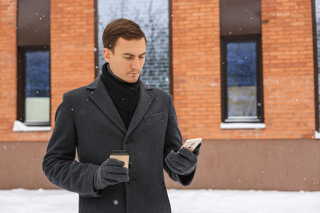 Serious businessman in winter coat surfing smartphone and having coffee break in city street