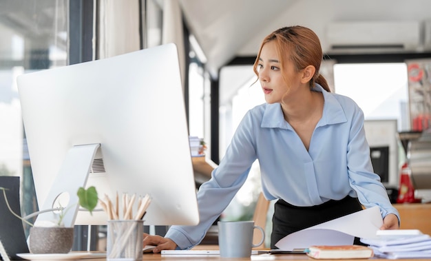 Serious businesswoman using desktop computer and holding papers preparing report analyzing work results female executive doing paperwork at workplace using computer online for data analysis