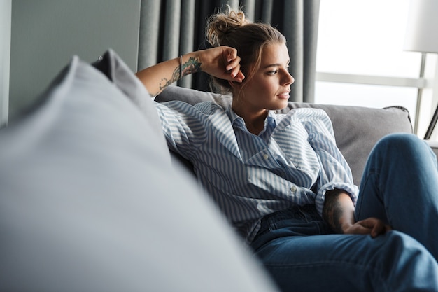 serious caucasian woman in striped shirt looking aside while sitting on sofa at living room