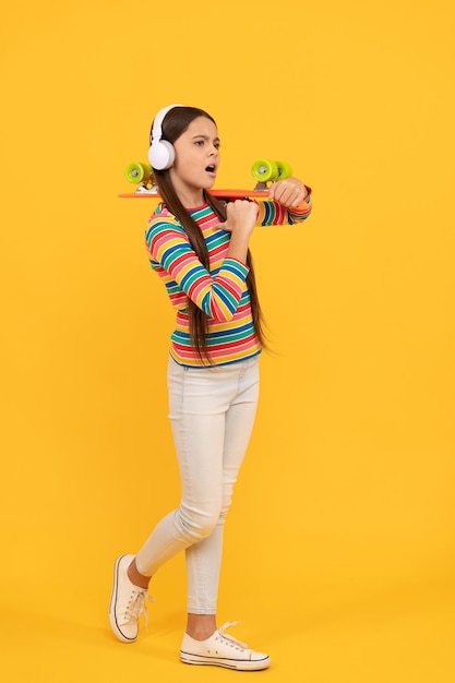 Serious child in headphones with penny board. teen girl listen music. childhood activity. hipster kid skateboarder in wireless earphones. penny skateboard. skateboarding.