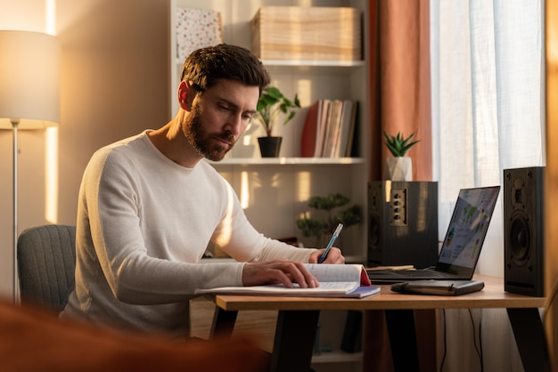 Serious concentrated bearded guy is laboring with pleasure at the laptop while writing something at the notebook