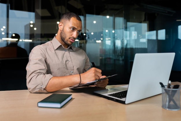 Serious concentrated businessman writing document while sitting in office using laptop for online