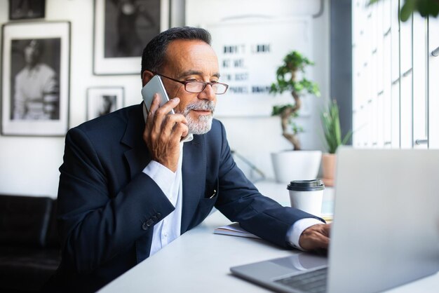 Serious concentrated busy old caucasian man in suit glasses calls by phone typing on laptop