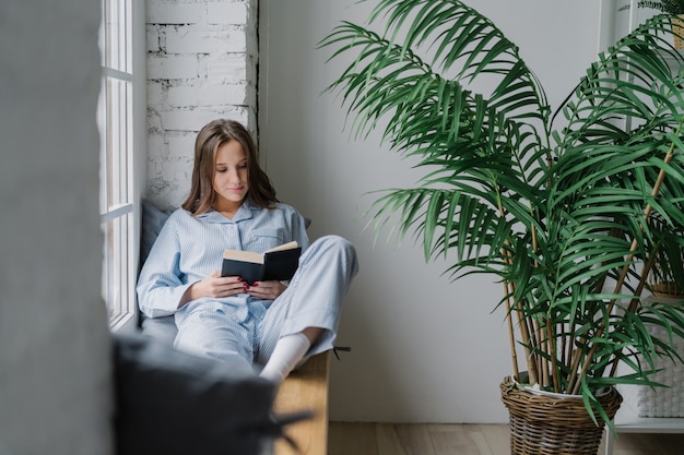 Serious concentrated female student focused into textbook, wears pyjamas, sits on window sill in cozy room