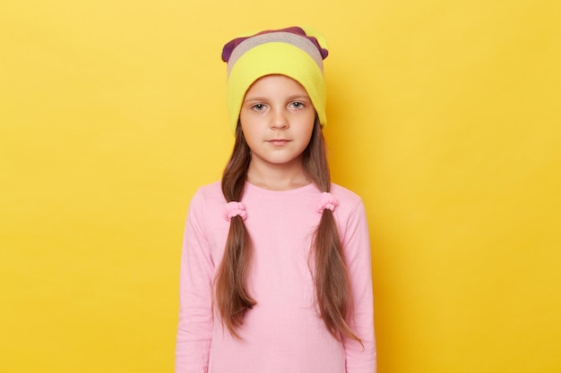 Serious confident little girl with ponytails wearing pink shirt and beanie hat posing isolated over yellow background looking at camera with displeased expression