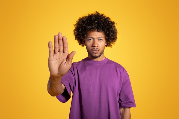 Serious confident mature black curly man in purple tshirt making stop gesture with hand