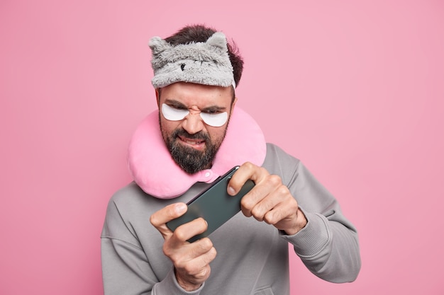 Serious displeased bearded man focused at smartphone plays video games online while traveling long distance in transport wears blindfold on forehead travel pillow around neck beauty patches under eyes