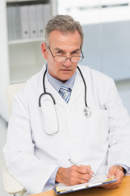 Serious doctor sitting at his desk writing on clipboard