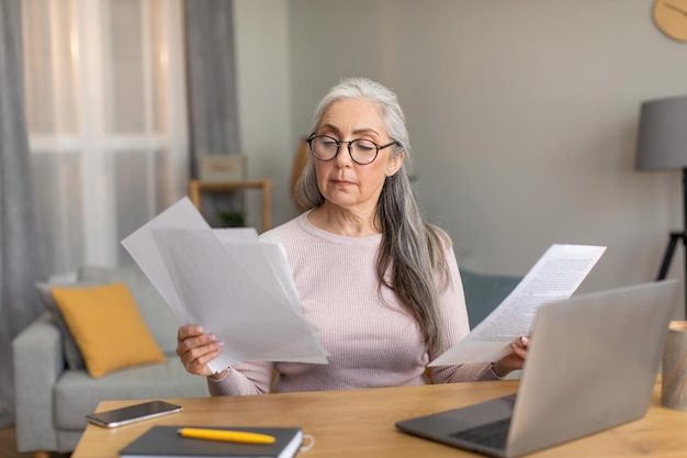 Serious european old female with gray hair in glasses works on computer and documents in room