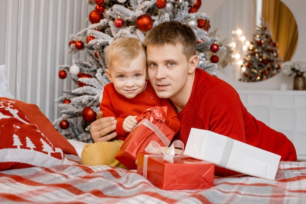 Serious father hugs his little child who opens Christmas gift and smile at camera with wrinkled forehead near Christmas tree. New Year Celebration.