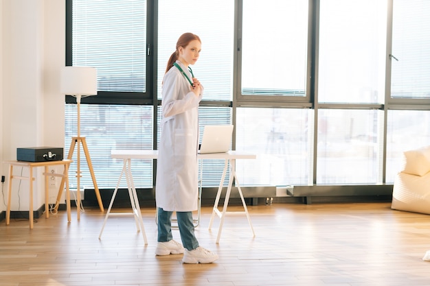 serious female doctor in white coat standing by window in sunny day in light medical clinic office