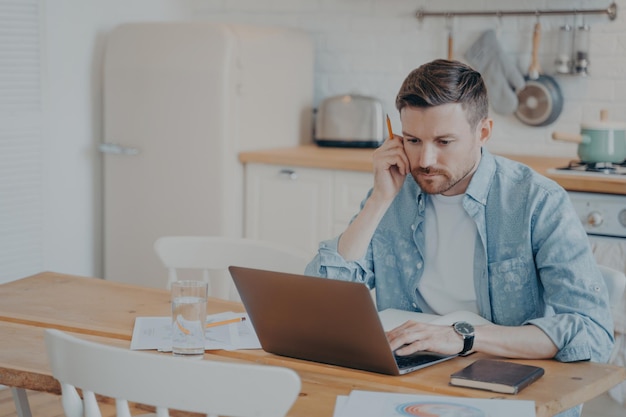 Serious focused bearded businessman in casual clothes working on laptop while sitting at kitchen table