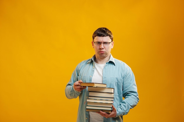 Serious focused man student in glasses holds stack of university books from college library on yellow background Education abroad concept Copy space