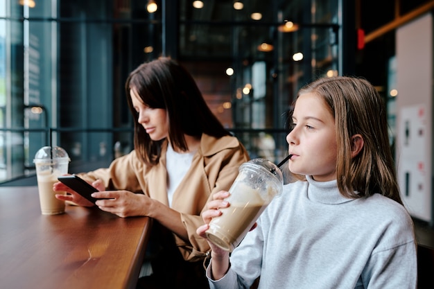 Serious girl drinking milk cocktail through straw and looking through window in cafe while her mother scrolling in gadget