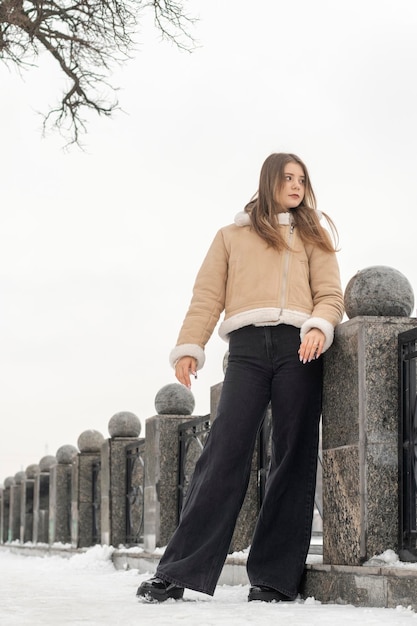 Serious girl in stylish black jeans and short beige sheepskin coat walks across bridge in park in winter