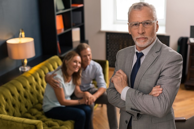 Serious grey-haired professional psychotherapist standing next to smiling patients in his office while looking at the camera