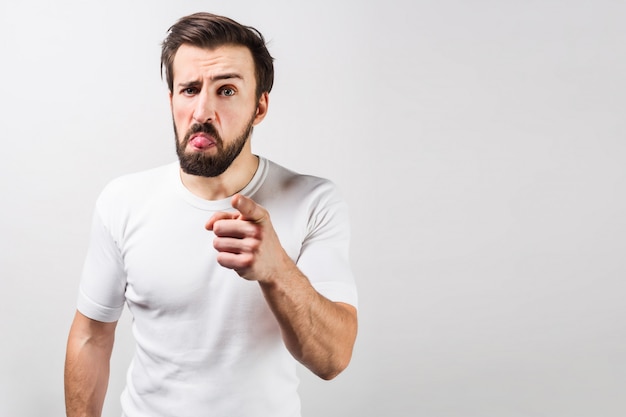 Serious guy in white shirt is standing at the left side of a frame and pointing straight forward. His face shows some strange emotions. Cut view. Isolated on white wall