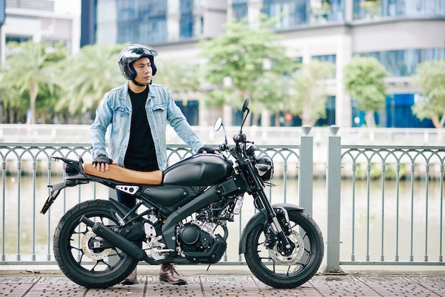 Serious handsome Asian man in helmet standing next to his motorcycle on river bank and looking away