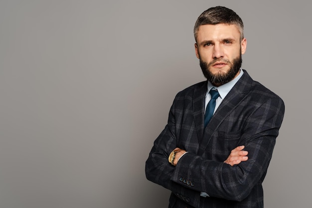 Serious handsome bearded businessman in suit with crossed arms on grey background