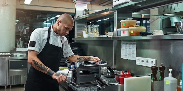 serious and handsome chef with tattoos on his hands rolling a black dough through pasta machine