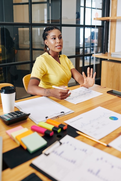 Serious Hispanic elegant business lady sitting at table in meeting room and telling her thoughts on project development