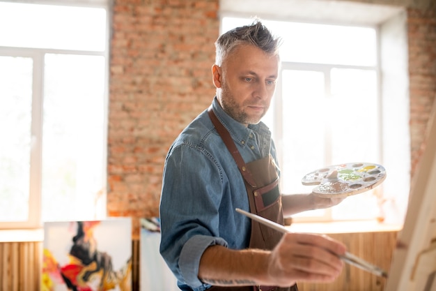 Serious inspired man with palette and paintbrush standing in front of easel and painting in workshop or studio