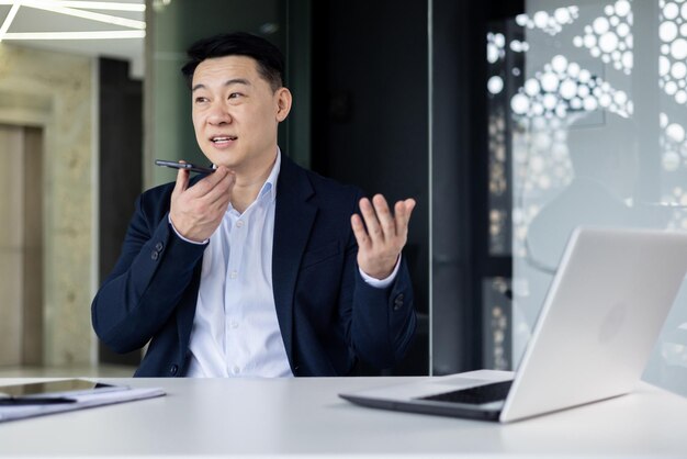 Serious korean man in a suit sits at a table in front of a laptop in a modern office and speaks on a