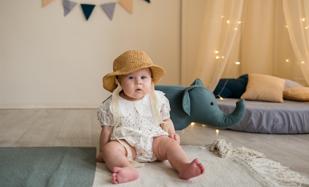 A serious little girl in a bodysuit and a straw hat is sitting on the floor in the nursery